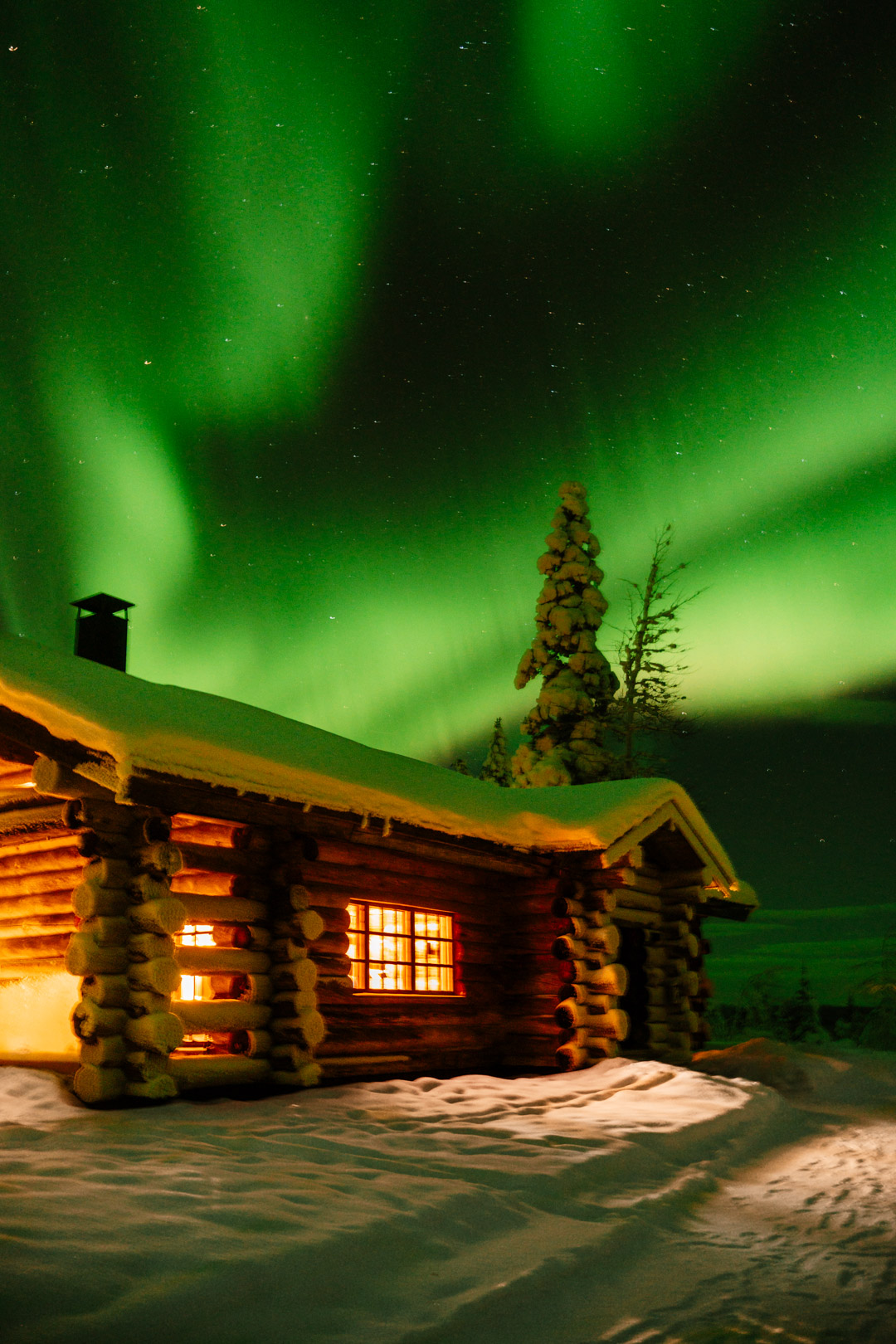 Chalet en rondins illuminé sous de grandes aurores boréales vertes, au milieu de la neige en Laponie finlandaise.