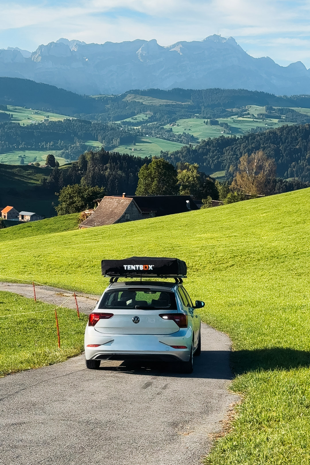 Voiture équipée d’une tente de toit TentBox repliée, sur une petite route traversant les prairies verdoyantes de Suisse, avec les montagnes en arrière-plan.